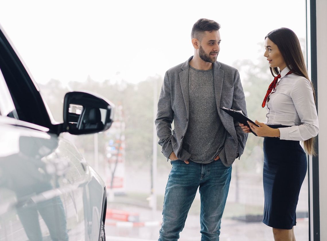 Atendente com prancheta conversa com um cliente ao lado de um carro, diante de uma janela ampla